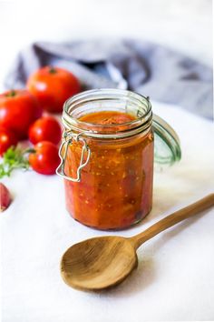 a jar filled with tomato sauce next to a wooden spoon and tomatoes on the table