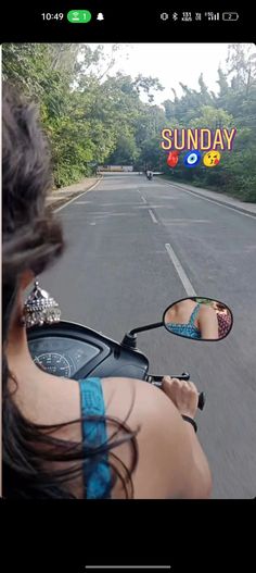 a woman riding a motorcycle down a street next to a lush green forest on a sunny day