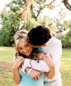 a man and woman hugging each other in front of a tree on the grass with trees in the background