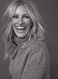 a smiling woman with long blonde hair wearing a gray sweater and turtle neck sweater, looking at the camera