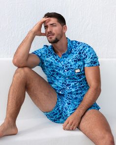 A man models the WET Stretch Shirt and matching beach shorts, sitting against a white textured wall. The vibrant blue outfit features a water-inspired pattern, designed for both comfort and fashion, perfect for resort or beachwear. Designed by DC2 Miami, a premier men's beachwear brand from Miami. Casual Short Sleeve Swimwear With Upf 50+, Gym Tank Tops, Tailored Shorts, Swim Brief, Men's Button Down Shirt, Beach Tops, Jogger Shorts, Gym Shirts, Sweat Proof