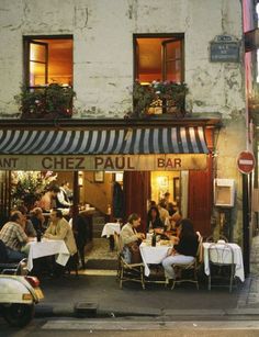 people sitting at tables in front of a restaurant on the side of a street with scooters parked nearby