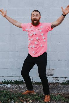 a man with his arms wide open standing in front of a white brick wall wearing a pink shirt and black pants