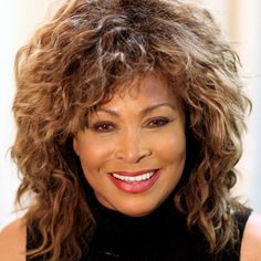 a woman with curly hair wearing a black top and smiling at the camera while standing in front of a curtain