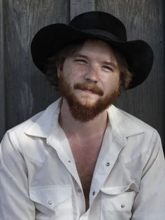 a man with a beard wearing a white shirt and black hat sitting in front of a wooden wall