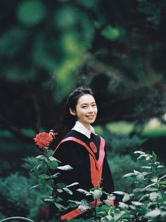 a woman standing in the woods holding onto a red flower and smiling at the camera