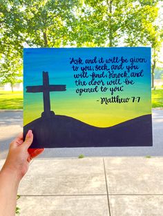 a hand holding up a card with a cross and bible verse written on it in front of trees