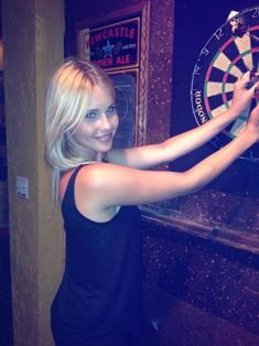 a beautiful blond woman standing in front of a dart board and holding a dartstick