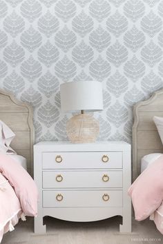 two white nightstands with pink pillows and blankets on them in front of a wallpapered bedroom