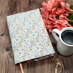 a cup of coffee sitting next to a notebook on top of a wooden table with flowers