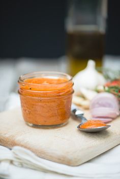 a wooden cutting board topped with a jar filled with orange sauce and vegetables next to a spoon