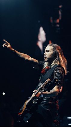 a man with long hair holding a guitar in his right hand and pointing at something