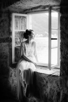 black and white photograph of woman sitting on window sill looking out at the ocean