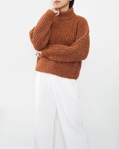 a woman in an orange sweater and white pants stands with her hands on her chin