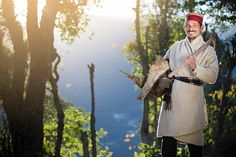 an old man holding a dead animal in the middle of a forest with trees and mountains in the background