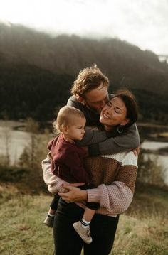 a woman holding a baby while standing on top of a grass covered field with mountains in the background