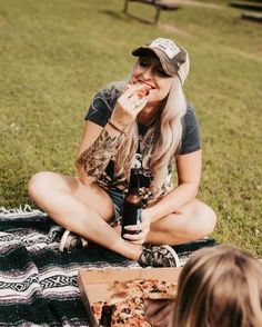 a woman sitting on the ground eating pizza and drinking beer while another person sits next to her