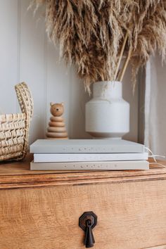 a stack of books sitting on top of a wooden dresser next to a vase filled with dry grass