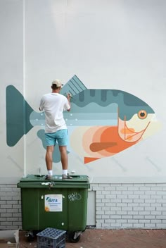 a man standing on top of a green trash can next to a fish painted on the side of a building