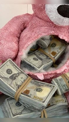 a pink teddy bear sitting on top of a pile of money with one hundred dollar bills in its mouth