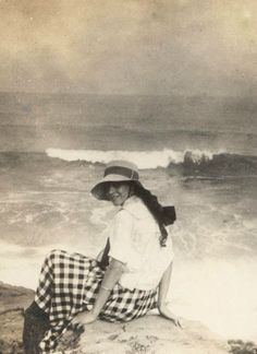 a woman sitting on top of a rock next to the ocean wearing a hat and checkered skirt