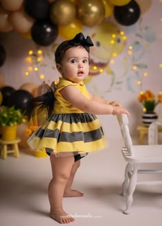 a baby girl in a yellow and black striped dress standing next to a white chair