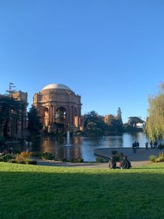 people are sitting on the grass in front of a pond and some buildings near by