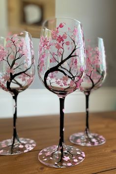 three wine glasses with pink flowers painted on them sitting on a wooden table in front of a mirror