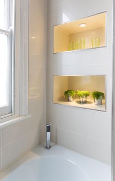 a bath tub sitting under a window next to a white tiled wall with shelves filled with potted plants