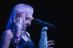 a woman with long pink hair holding a microphone and singing into a microphone in front of a black background