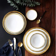 a table setting with white and gold plates, silverware and greenery on it