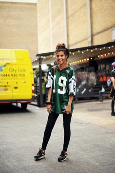 a woman in a football jersey is standing on the street with her legs crossed and smiling