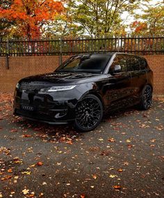 a black range rover parked in front of a brick wall with autumn leaves on the ground