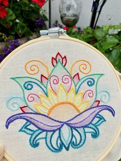 a hand is holding up a colorful embroidered flower on a white hoop with flowers in the background