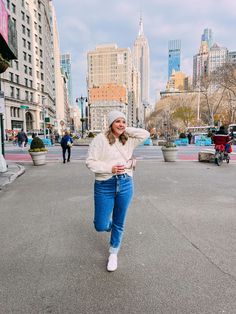 a woman is walking down the street with her hand on her head while wearing jeans and a white sweater