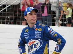 a man in a blue and yellow racing suit standing next to a fence with people watching from the bleachers