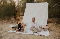 a man and woman sitting on a blanket in front of a white tarp with flowers