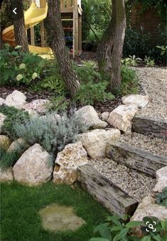 an outdoor play area with rocks, grass and trees in the foreground is a yellow slide