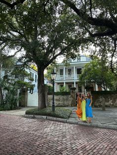 two women standing in front of a large tree on a brick road with houses behind them