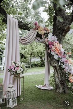 an outdoor wedding ceremony with pink drapes and flowers on the tree trunk, in front of a white gazebo