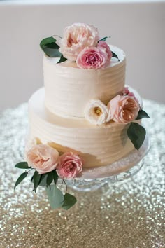 a three tiered cake with pink flowers and greenery sits on a gold sequin tablecloth