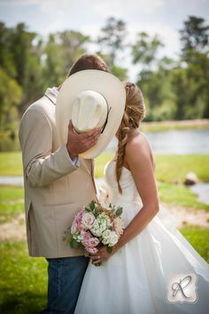 a bride and groom kissing in front of a pond with flowers on the grass, wearing cowboy hats