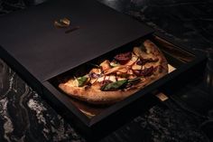 a pizza sitting inside of a black box on top of a marble countertop next to a knife and fork