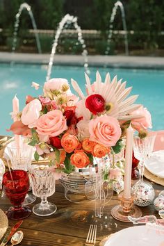 a table with flowers, candles and wine glasses on it near a pool in the background