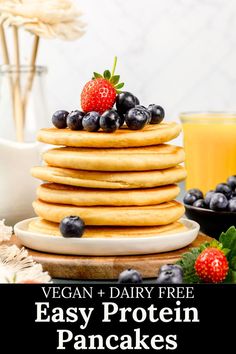 A stack of vegan protein pancakes are on a white plate on the kitchen table. Fresh blueberries and strawberries are on the plate. Juice and flowers are blurred in the background. Text on the image reads, "vegan and dairy free easy protein pancakes". Easy Protein Pancakes