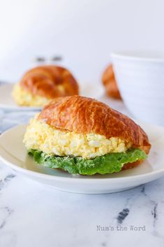 a croissant sandwich on a white plate next to a cup and bowl filled with eggs