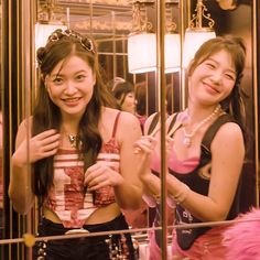 two young women standing next to each other in front of a mirror with pink feathers on it