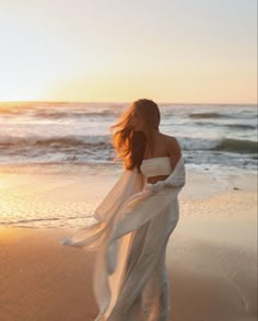 a woman is walking on the beach with her dress blowing in the wind as the sun sets