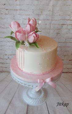 a white cake with pink flowers on top sitting on a table next to a brick wall