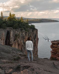 a woman standing on the edge of a cliff looking out at a boat in the water